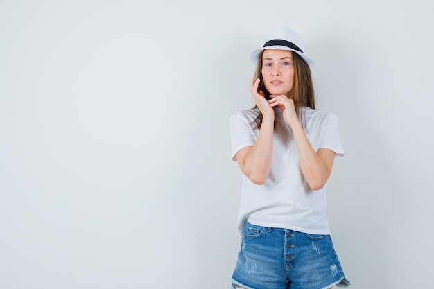 Jonge vrouw wat betreft haar gezichtshuid in wit t-shirt, korte broek, hoed en elegant op zoek.