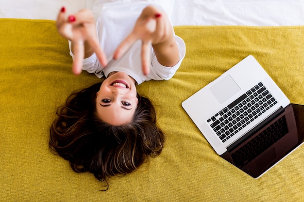 Jonge vrouw wees overwinning teken liggend in bed met laptop van bovenaf. Bovenaanzicht.