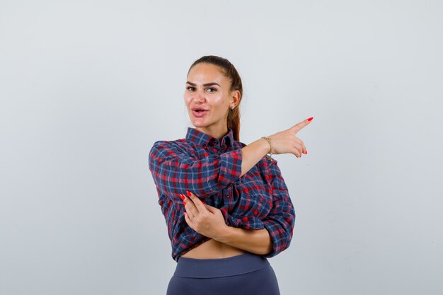 Jonge vrouw wijst naar de rechterkant in geruit hemd, broek en ziet er zelfverzekerd uit. vooraanzicht.