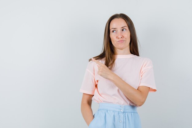 Jonge vrouw wijst naar kant in t-shirt, rok en op zoek sluw. vooraanzicht.