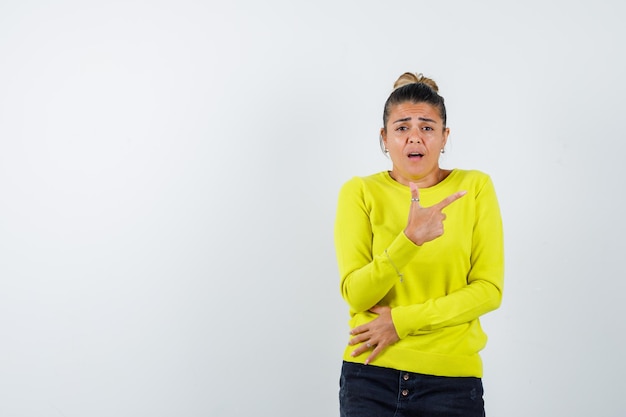 Jonge vrouw wijst naar rechts met wijsvinger in gele trui en zwarte broek en kijkt gehaast