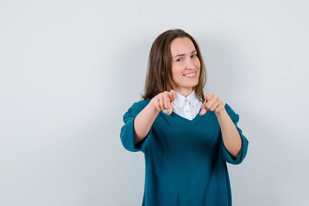 Jonge vrouw wijst naar voren in trui over shirt en ziet er vrolijk uit. vooraanzicht.