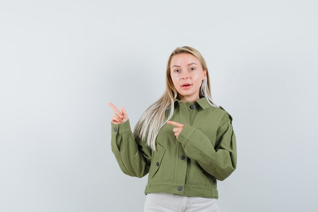 Jonge vrouw wijst opzij in groene jas, spijkerbroek, vooraanzicht.