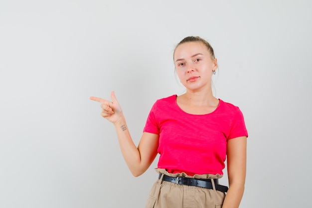 Jonge vrouw wijst opzij in t-shirt, broek en kijkt vrolijk, vooraanzicht.