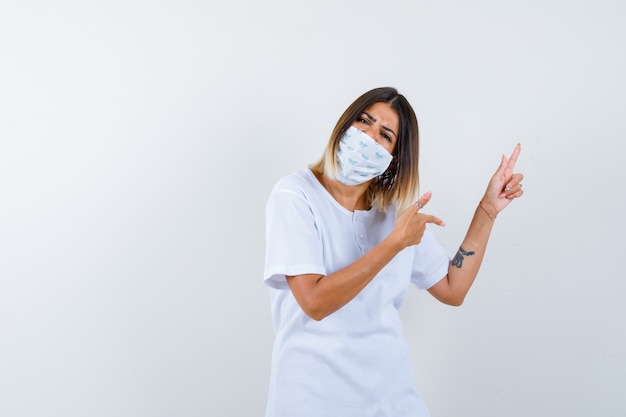 Jonge vrouw wijst opzij in t-shirt, masker en kijkt aarzelend, vooraanzicht.