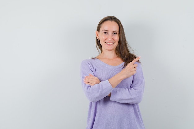 Jonge vrouw wijst opzij terwijl ze haar armen kruist in een lila blouse en er tevreden uitziet. vooraanzicht.