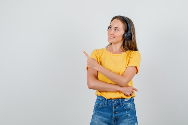 Jonge vrouw wijst weg in t-shirt, korte broek, koptelefoon en kijkt blij