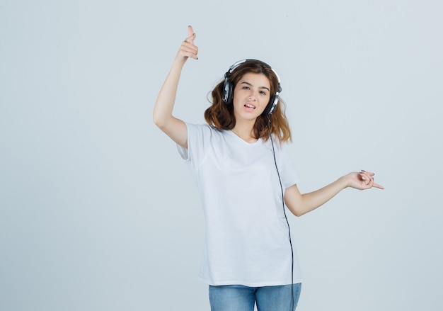 Jonge vrouw wijst weg terwijl u geniet van muziek met koptelefoon in wit t-shirt, spijkerbroek en op zoek gelukkig, vooraanzicht.