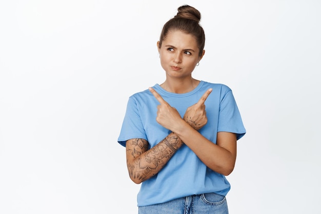 Jonge vrouw wijst zijwaarts met de vingers en kijkt naar links, maakt haar keuze tussen twee opties, staande in blauwe tshirt op witte achtergrond.