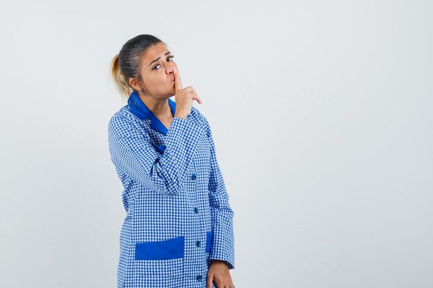 Jonge vrouw wijsvinger op mond zetten, stiltegebaar in blauw geruit pyjama shirt tonen en er mooi uitzien, vooraanzicht.