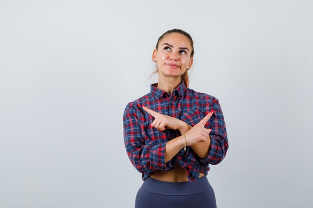 Jonge vrouw wijzend naar beide hoeken in geruit hemd, broek en aarzelend kijkend, vooraanzicht.