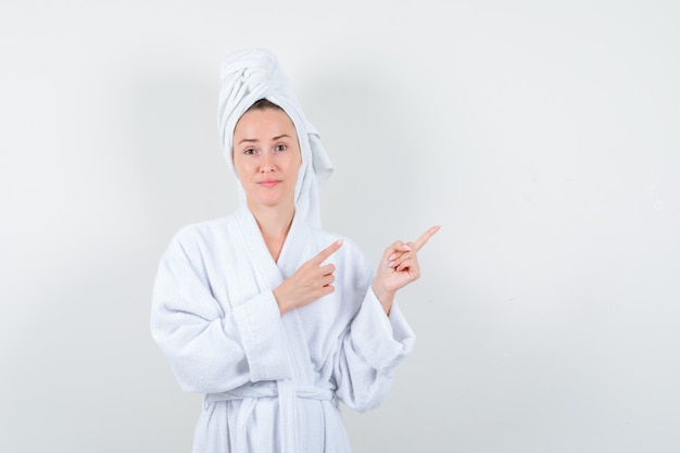 Jonge vrouw wijzend op de rechterbovenhoek in witte badjas, handdoek en op zoek verstandig, vooraanzicht.