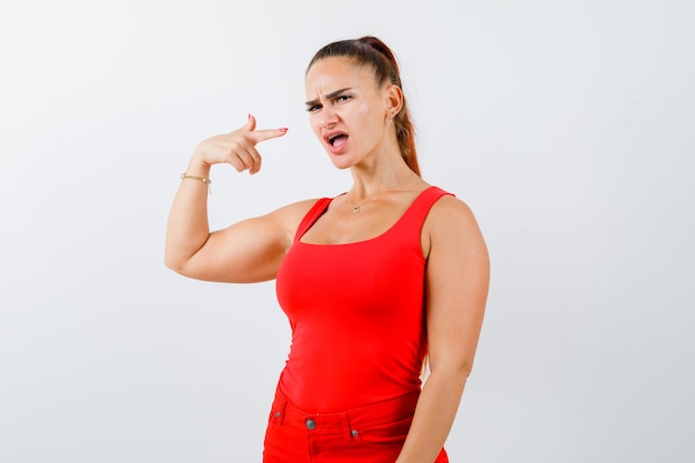 Jonge vrouw wijzend op zichzelf in rode tank top, broek en aarzelend, vooraanzicht op zoek.