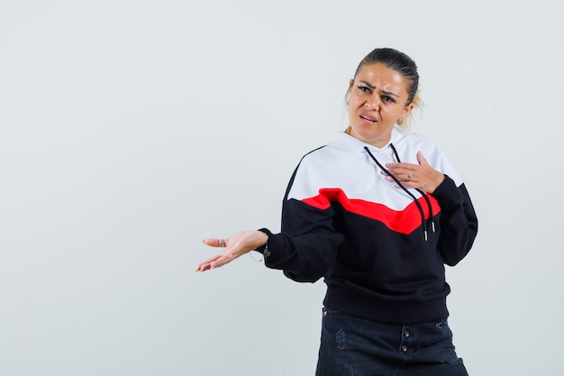 Jonge vrouw wijzend op zichzelf terwijl ze open palm opzij spreidt in kleurrijke sweater en agressief, vooraanzicht kijkt.