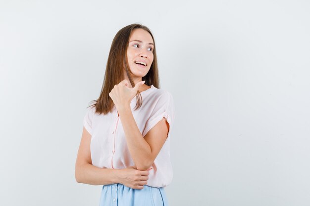 Jonge vrouw wijzende duim terug in t-shirt, rok en op zoek optimistisch, vooraanzicht.