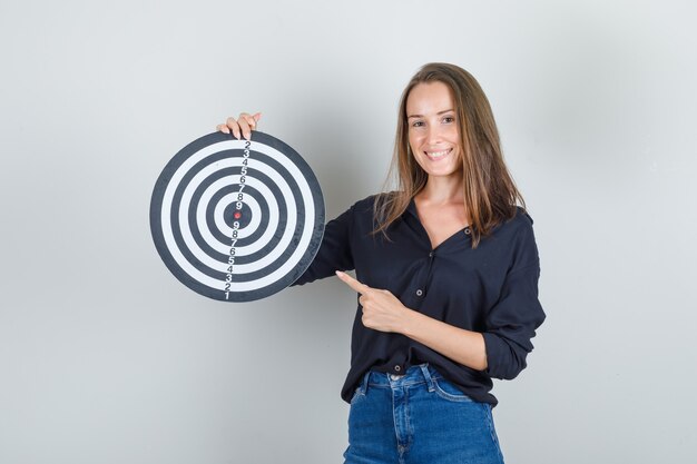 Jonge vrouw wijzende vinger op dartbord in zwart shirt, jeans broek en op zoek vrolijk