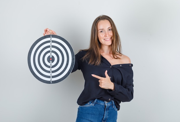 Jonge vrouw wijzende vinger op dartbord in zwart shirt, jeans broek en op zoek vrolijk