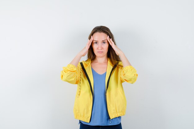 Jonge vrouw wrijft over haar slapen in t-shirt, jas en ziet er vermoeid uit, vooraanzicht.