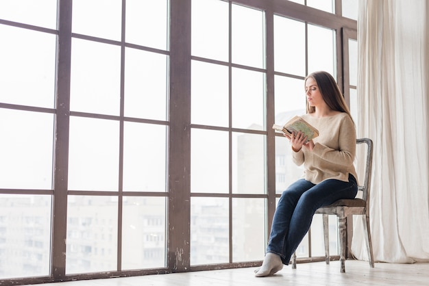 Jonge vrouw zitten in de buurt van het venster het lezen van het boek