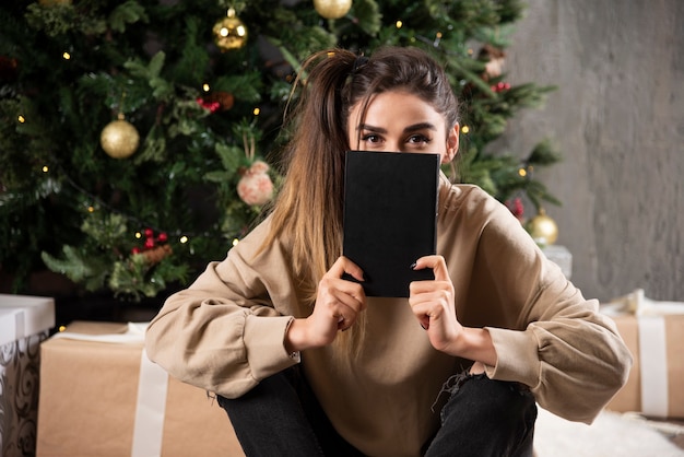 Jonge vrouw zitten met zwarte notebook in de buurt van kerstboom.