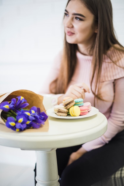 Gratis foto jonge vrouw zittend aan witte tafel met bloemboeket en bitterkoekjes op plaat