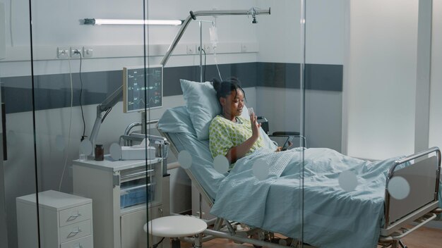 Jonge vrouw zwaaien op video-oproep op smartphone in ziekenhuisafdeling bed. Patiënt met IV-druppelzak en oximeter die online conferentie op afstand op internet gebruikt om met vrienden te praten en te herstellen