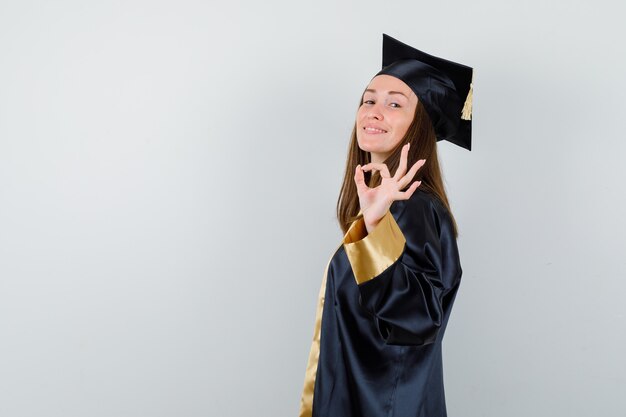 Jonge vrouwelijke afgestudeerde die ok gebaar in academische kleding toont en positief, vooraanzicht kijkt.