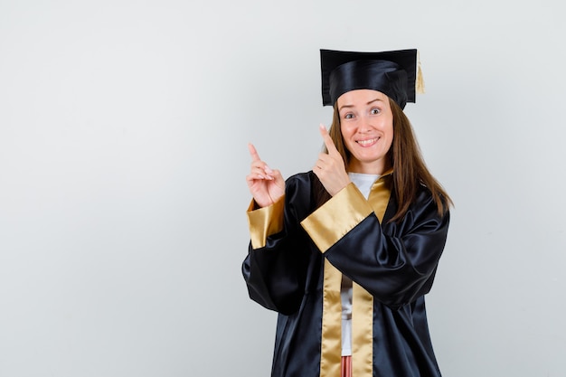 Jonge vrouwelijke afgestudeerde die omhoog wijst in academische kleding en er vrolijk uitziet. vooraanzicht.