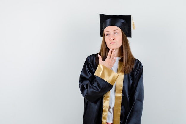 Jonge vrouwelijke afgestudeerde die zich in academische kleding bevindt en peinzend kijkt. vooraanzicht.