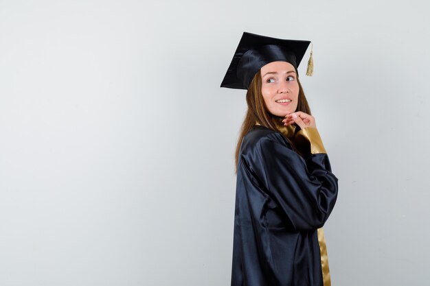 Jonge vrouwelijke afgestudeerde kijkt over de schouder in academische kleding en ziet er prachtig uit. vooraanzicht.