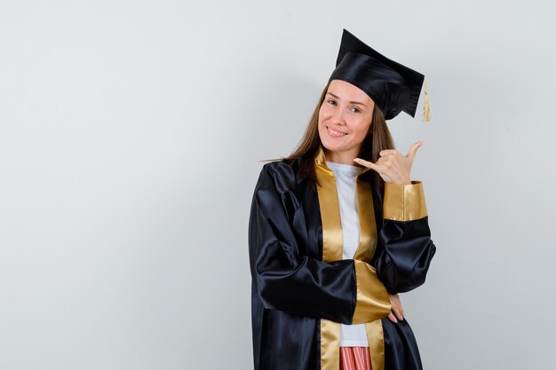 Jonge vrouwelijke afgestudeerde telefoongebaar in academische kleding tonen en op zoek vrolijk. vooraanzicht.