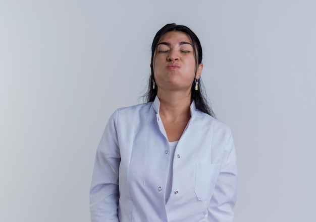 Jonge vrouwelijke arts die medische mantel puffende wangen met gesloten geïsoleerde ogen draagt