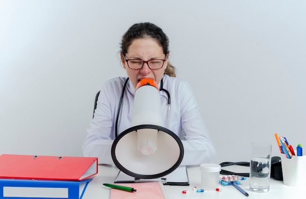 Jonge vrouwelijke arts het dragen van medische mantel en stethoscoop en bril zit aan bureau met medische hulpmiddelen schreeuwen in luidspreker met gesloten ogen geïsoleerd