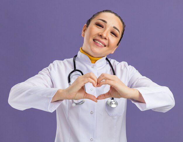 Jonge vrouwelijke arts in een witte medicijnjas met een stethoscoop om de nek die een hartgebaar maakt met vingers die vrolijk glimlachen over de paarse muur