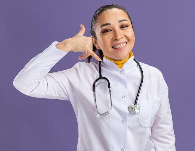 Jonge vrouwelijke arts in een witte medicijnjas met een stethoscoop om de nek met een glimlach op het gezicht die me een gebaar maakt dat over de paarse muur staat