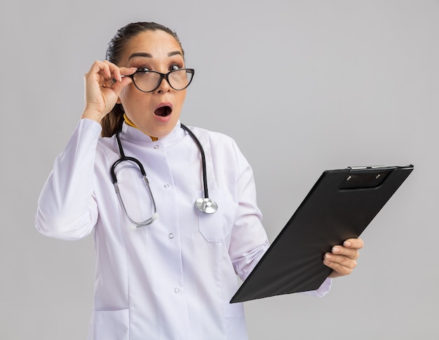 Jonge vrouwelijke arts in een witte medische jas met een bril met een stethoscoop om de nek met een klembord verrast