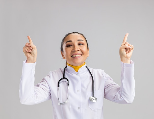 Jonge vrouwelijke arts in een witte medische jas met een stethoscoop om de nek, blij en vrolijk met wijsvingers die over een witte muur staan