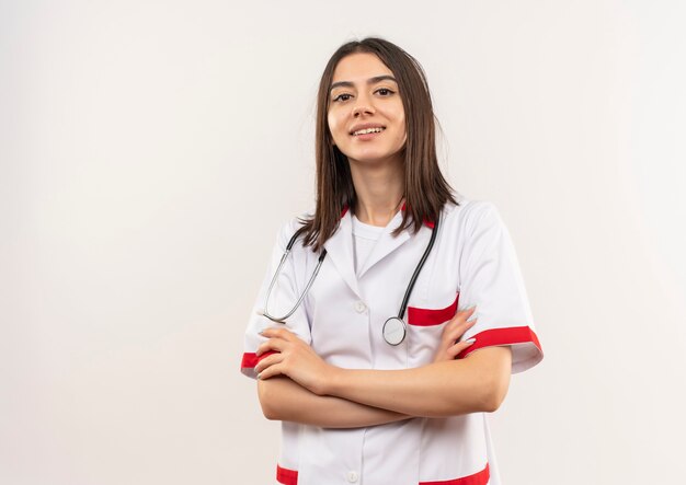 Jonge vrouwelijke arts in witte jas die met een stethoscoop om haar hals naar voren kijkt met hand op borst die zelfverzekerd over witte muur kijkt