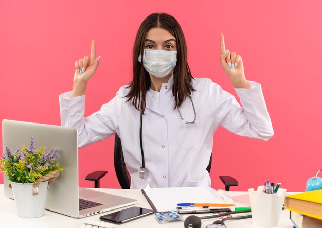 Jonge vrouwelijke arts in witte jas met beschermend gezichtsmasker en stethoscoop om haar nek omhoog met wijsvingers zittend aan tafel met laptop over roze muur