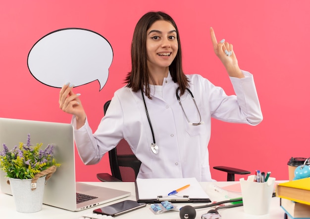 Jonge vrouwelijke arts in witte jas met een stethoscoop om haar nek die een leeg teken van de tekstballon houdt die met vinger omhoog wijst aan de tafel met laptop over roze muur