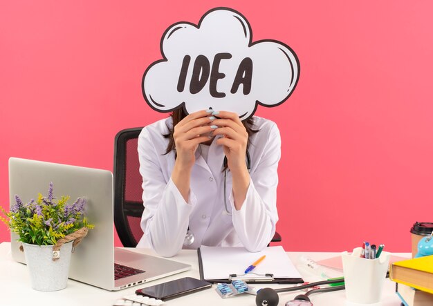 Jonge vrouwelijke arts in witte jas met een stethoscoop om haar nek die haar gezicht bedekt met het idee van het toespraakbel teken aan de tafel zitten met laptop over roze muur