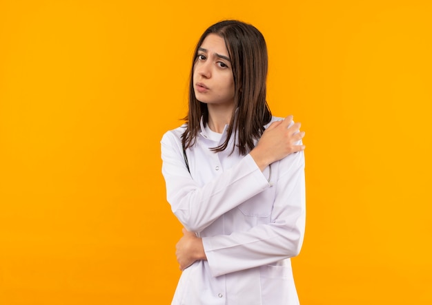 Jonge vrouwelijke arts in witte jas met een stethoscoop om haar nek kijkt onwel wat betreft haar schouder met pijn die zich over oranje muur bevindt
