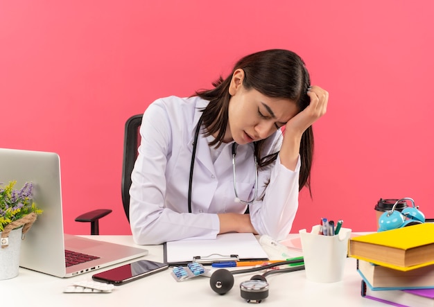 Jonge vrouwelijke arts in witte jas met een stethoscoop om haar nek op zoek naar band en overwerkt zittend aan tafel met laptop over roze muur