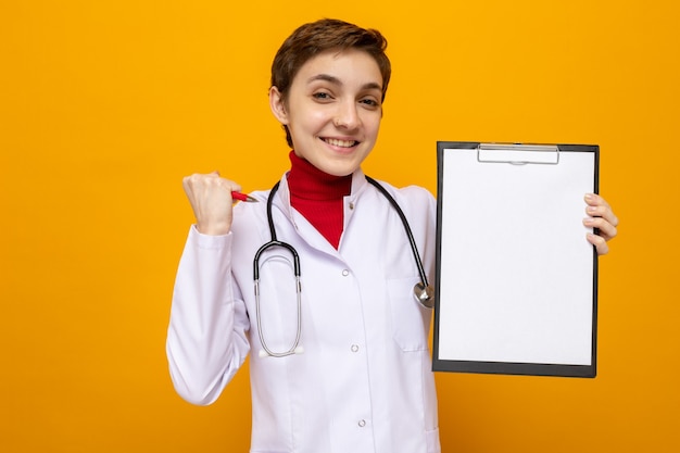 Jonge vrouwelijke arts in witte jas met stethoscoop die klembord vasthoudt met blanco pagina's die er blij en opgewonden uitzien met gebalde vuist Gratis Foto