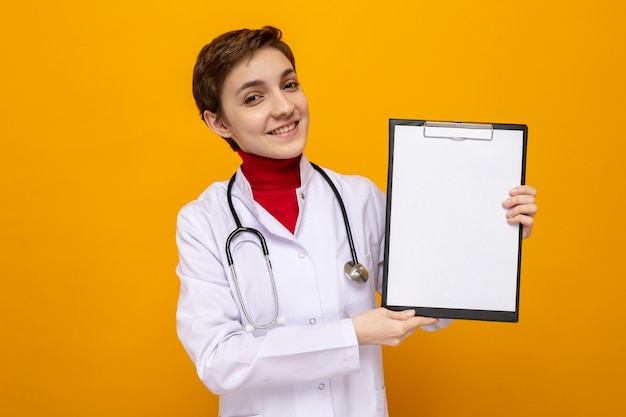 Jonge vrouwelijke arts in witte jas met stethoscoop die klembord vasthoudt met blanco pagina's die er gelukkig en positief uitzien en vrolijk glimlachen