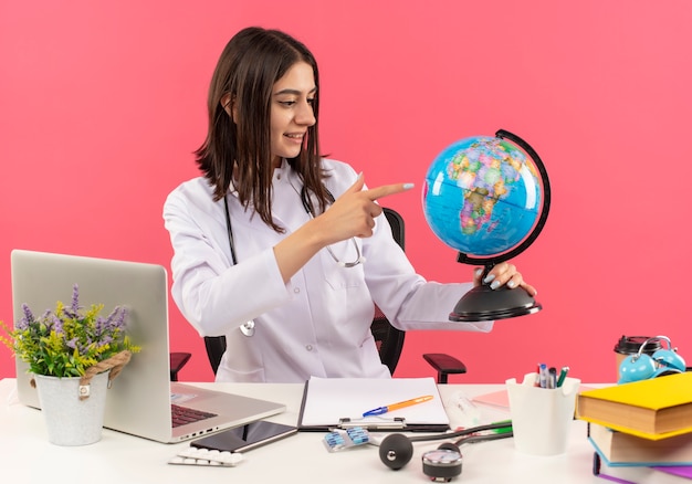Jonge vrouwelijke arts in witte jas met stethoscoop om haar nek die bol houdt die met glimlach op gezicht kijkt zittend aan tafel met laptop over roze muur