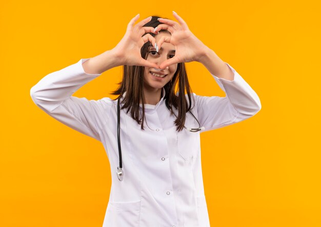 Jonge vrouwelijke arts in witte jas met stethoscoop om haar nek die hartgebaar maakt met vingers die naar voren kijken met glimlach op gezicht die zich over oranje muur bevinden