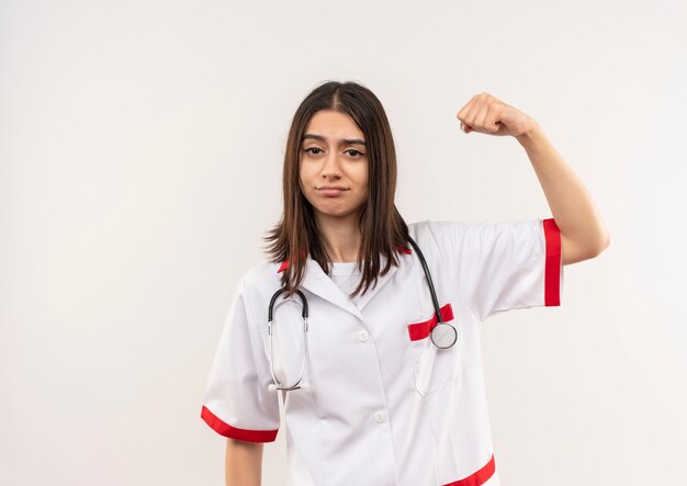 Jonge vrouwelijke arts in witte jas met stethoscoop om haar nek gebalde vuist hand opsteken op zoek zelfverzekerd, winnaar concept staande over witte muur