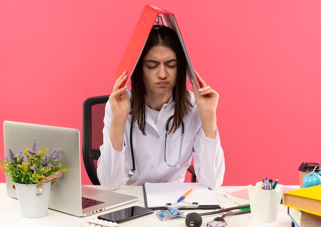 Jonge vrouwelijke arts in witte jas met stethoscoop om haar nek hoding map boven haar hoofd op zoek moe en overwerkt zittend aan tafel met laptop over roze muur Gratis Foto