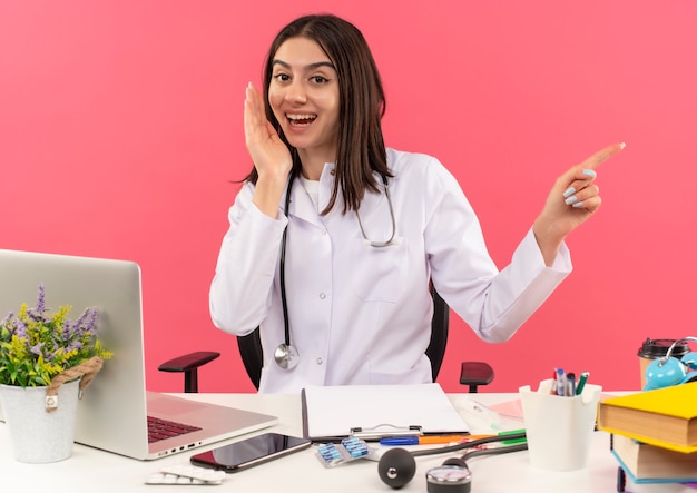 Jonge vrouwelijke arts in witte jas met stethoscoop om haar nek naar voren kijkend glimlachend met blij gezicht wijzend met vinger naar de zijkant zittend aan tafel met laptop over roze muur